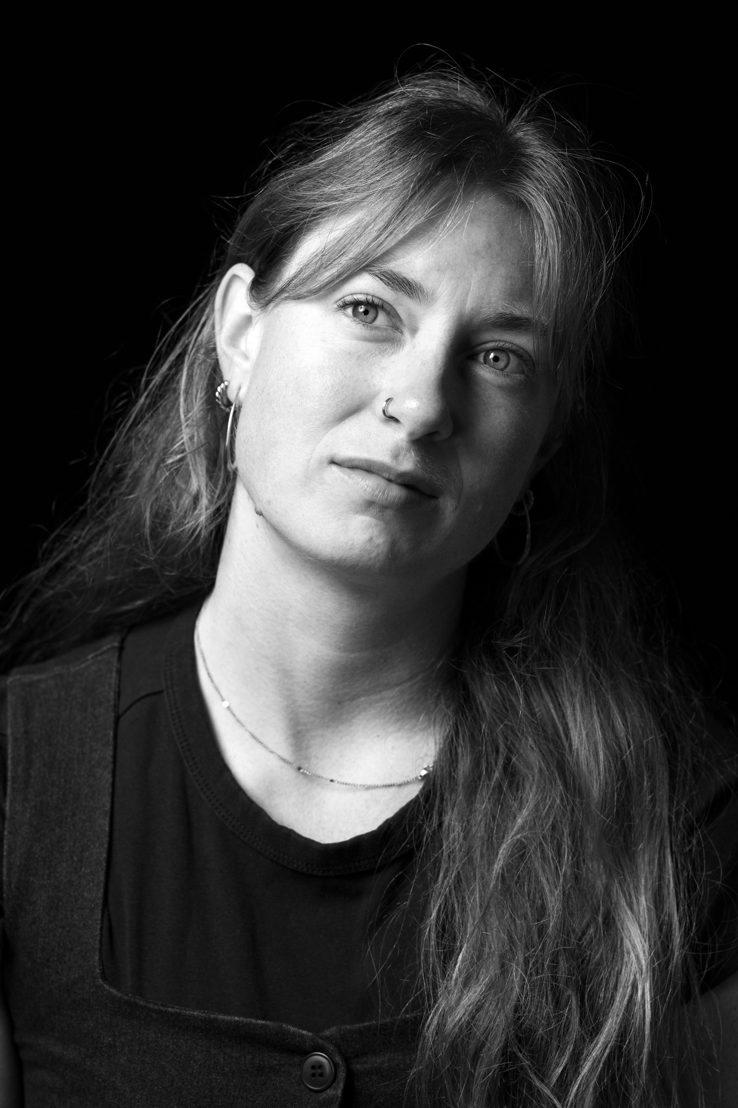 Black-and-white close-up portrait of a woman with long hair, nose ring, and hoop earrings, looking slightly to the side against a dark background.