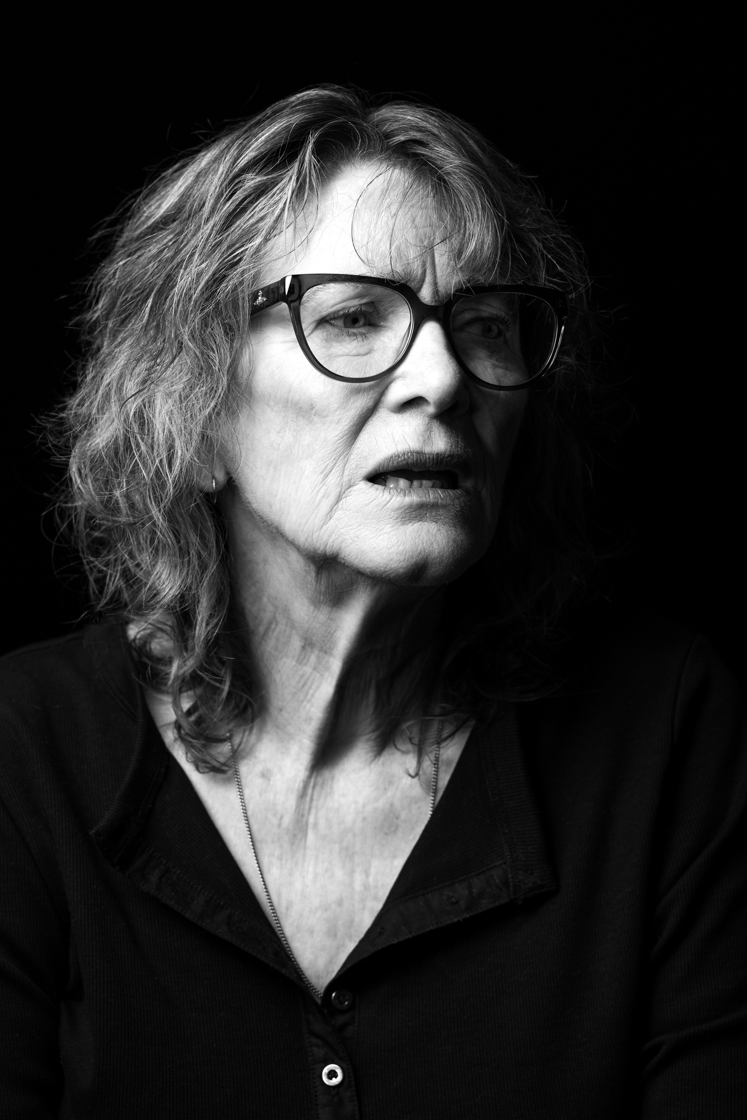 Older woman with wavy hair and large black-framed glasses, facing right against a dark background.
