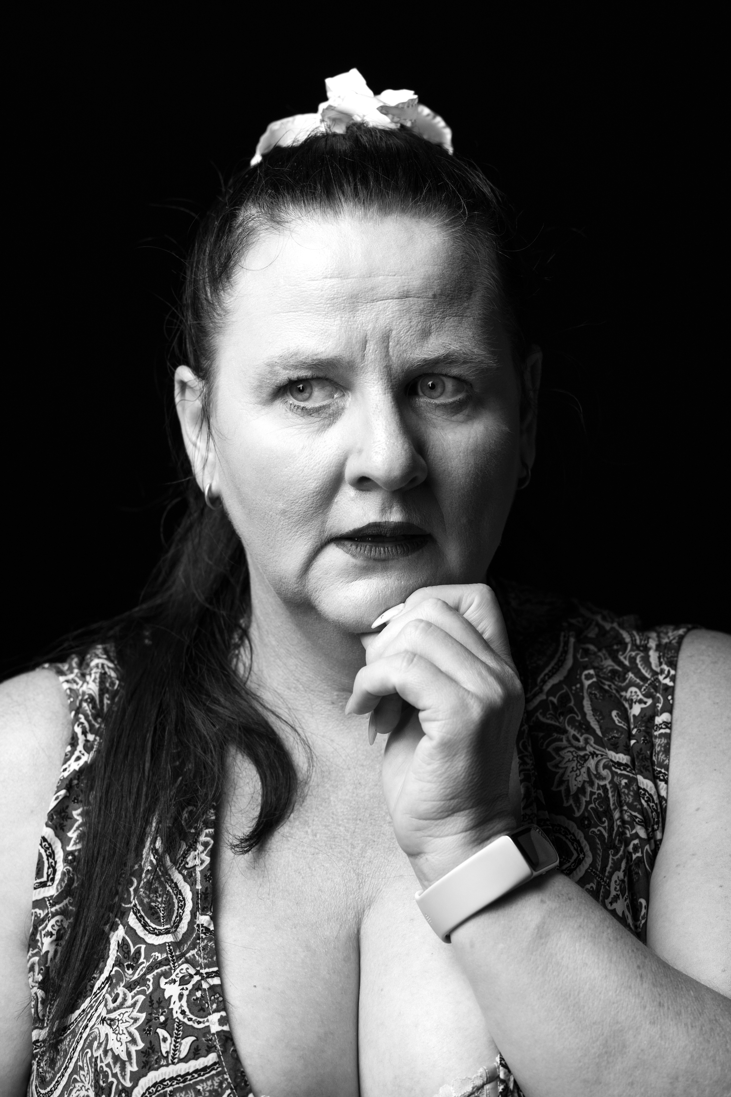 Black-and-white portrait of a woman with a thoughtful expression, chin resting on clasped hands, wearing a paisley-pattern top and a light smartwatch on her wrist with a scrunchie in her hair.