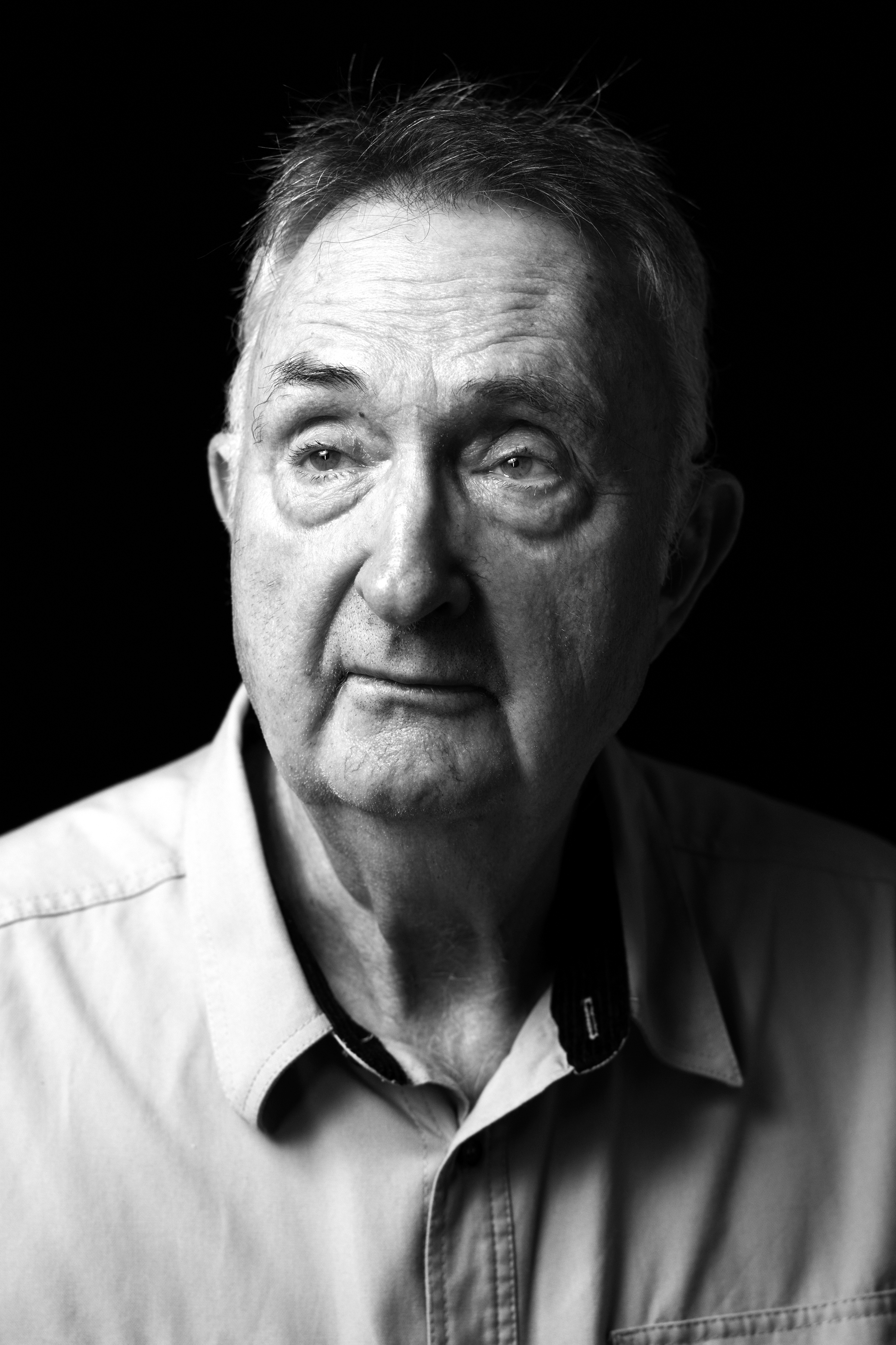 Black-and-white close-up portrait of an older man, showing his eyes and part of his face and shirt collar against a dark background.