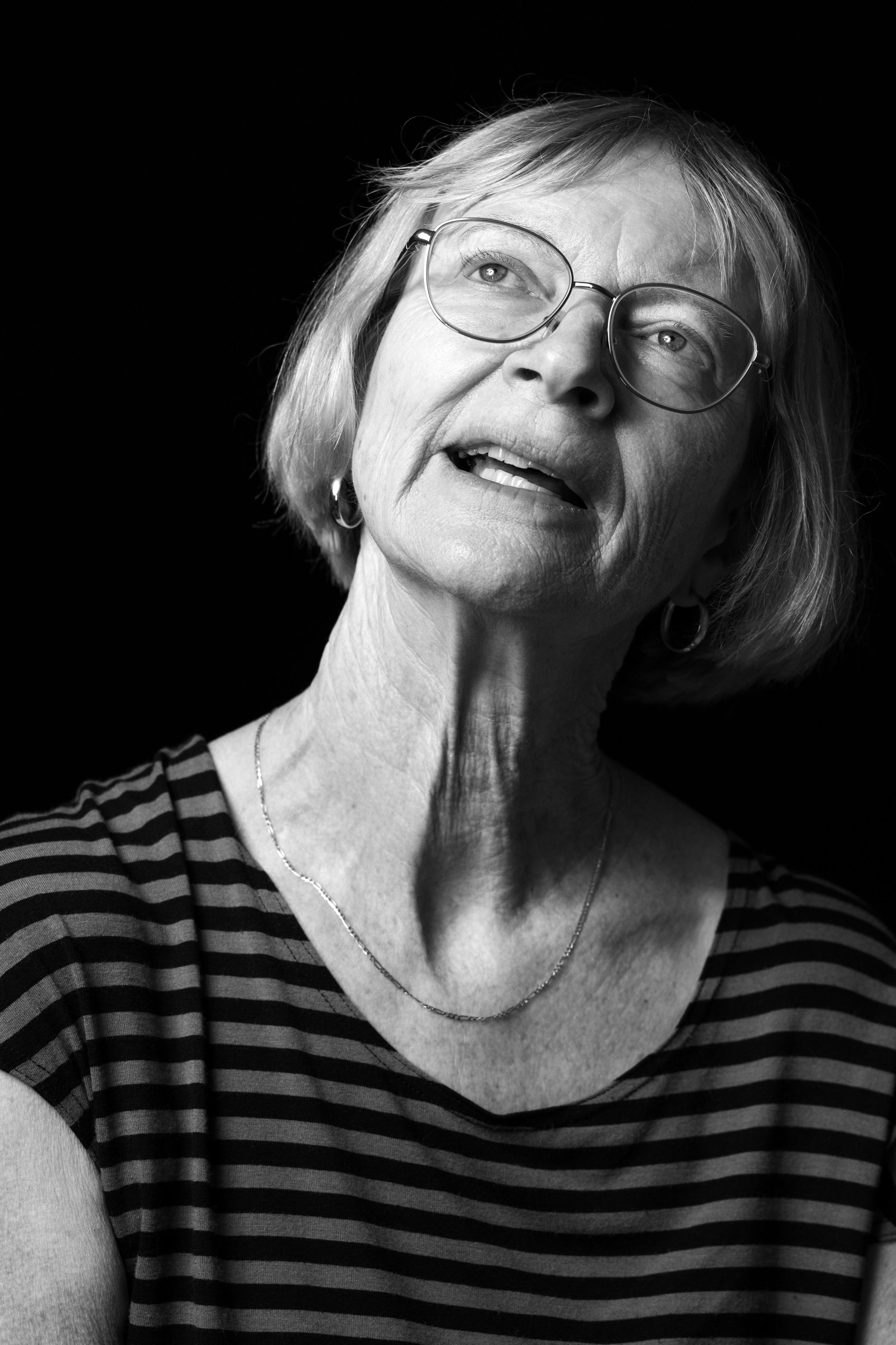 Close-up portrait of an older woman wearing round glasses, smiling, in a black-and-white photo with a dark background.